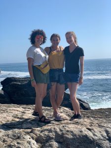 three women stand on a large rock with the blue ocean behind them